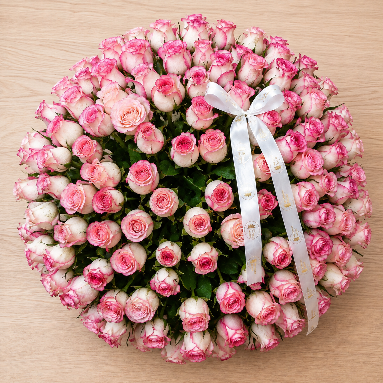 A large round floral arrangement of pink and white roses with green foliage, tied with a white satin ribbon, photographed from above on a light wood surface.