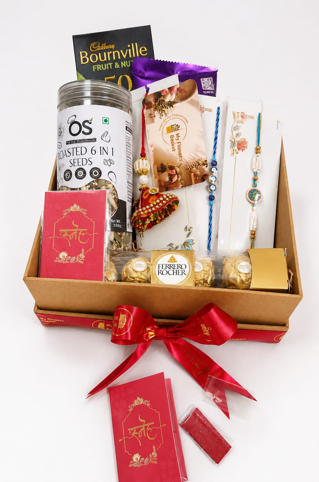 A curated gift basket in a kraft box filled with assorted packaged snacks and sweets, tied with a red ribbon. Visible items include a jar of roasted seeds, a Cadbury Bournville fruit & milk chocolate bar, Ferrero Rocher chocolates, and red decorative cards with gold script.
