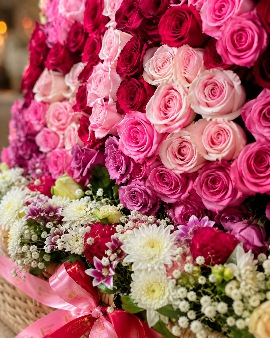 A large, vibrant floral arrangement of roses and chrysanthemums in shades of pink, magenta, white, and green, tied with a pink ribbon and presented in a basket.
