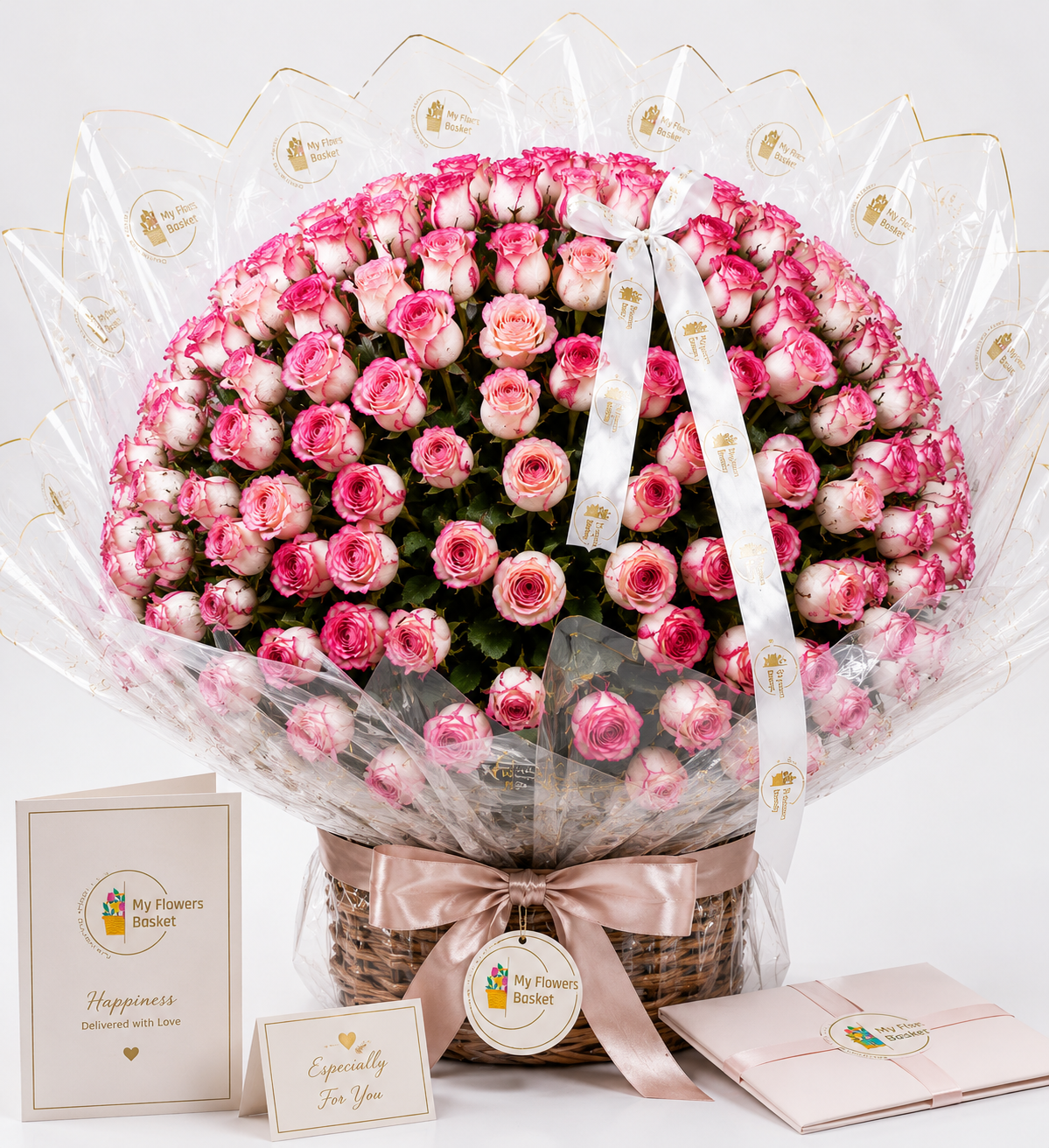 A large bouquet of pink roses arranged in a round shape inside a wicker basket, wrapped in clear cellophane with a white ribbon printed with gold hearts. Two pink greeting cards are placed in front on a white background.
